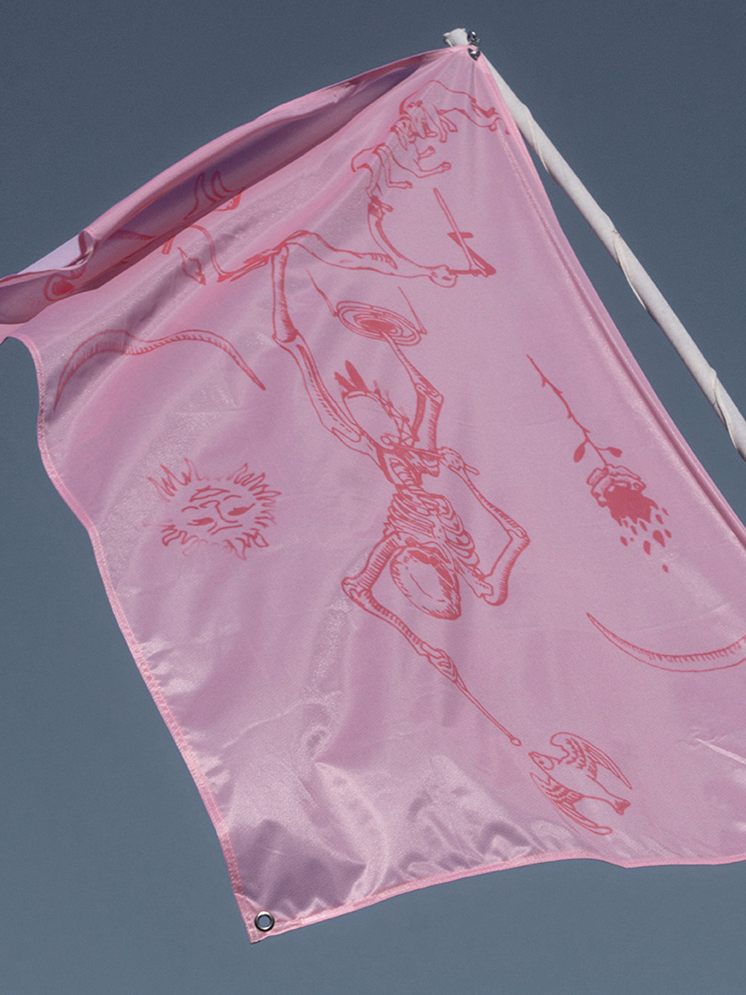 A pink flag with a red print. The print shows a quote from Holbeins' Images of Death, with the drumming skeleton wearing a prosthetic leg. There is also a burning rose, a laughing sun, a quotation from Master E.S. of two dogs wrestling over a bone, a flying fish and an unidentifiable quadruped with offspring.